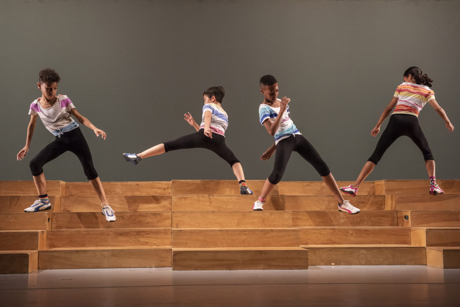 Four young dancers wearing colorful t-shirts and black leggings perform dynamic moves on a wooden staircase. The dancers are positioned in varied energetic stances, showcasing synchronized choreography against a simple backdrop.